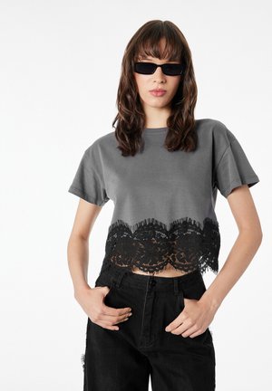 Woman wearing black sunglasses, a gray cropped t-shirt with black lace trim, and high-waist black pants with hands in pockets.