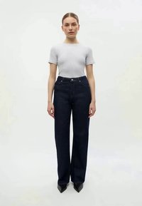 Botas de ganga azul-escura de perna reta com cinco bolsos, fecho de botão e zipper, e detalhes de costura em contraste, combinadas com uma t-shirt justa cinza-claro.