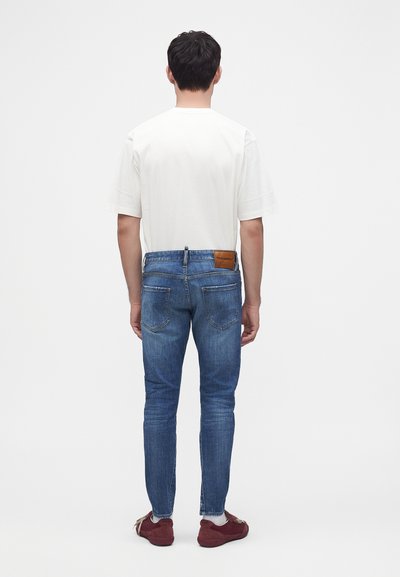 Homme vu de dos portant un t-shirt blanc uni, un jean slim bleu avec une étiquette en cuir de la marque et des baskets rouges avec des chaussettes blanches.