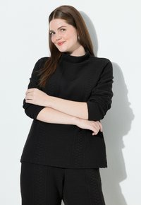 Młoda kobieta z długimi brązowymi włosami stoi z założonymi rękami, ubrana w fakturowany czarny sweter i dopasowane spodnie na białym tle.
