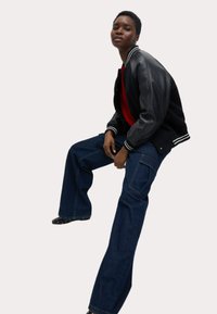 Chaqueta bomber negra con mangas de cuero, combinada con unos pantalones vaqueros oscuros y holgados. Suéter rojo visible debajo. Zapatos negros con lunares.