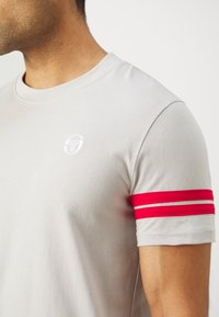 Grå bomulls t-skjorte med rund hals, som har en liten hvit logo og to røde striper på ermet for aksent. Glatt tekstur.