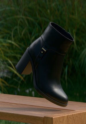 Stivaletto alla caviglia in pelle nera con tacco a blocco e zip laterale, mostrato sopra una superficie in legno con sfondo sfocato di fogliame verde.