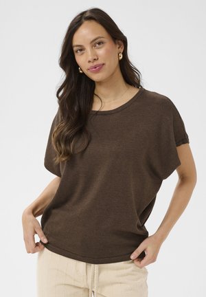 Pikkade tumepruunide juustega naine, kellel on kuldsed rõngaskõrvarõngad, pruun lühikeste varrukatega top ja kreemikad kudumispüksid, seisab ühevärvilise heleda tausta ees.