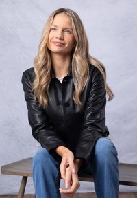 Black leather jacket with a collared design and button closure, worn over a white top. Model seated, paired with blue jeans. Smooth texture.
