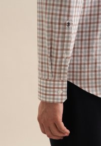 Camicia a scacchi marrone e bianca a maniche lunghe, con polsini a bottone e un piccolo dettaglio ricamato sulla manica. Tessuto di cotone.
