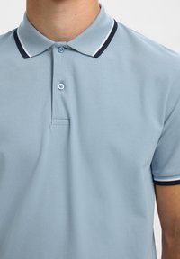 Homme portant un polo bleu clair avec un col et des manches rayés bleu marine et blanc, deux boutons fermés au niveau du cou.