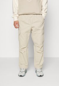 Pantaloni cargo beige con vita elastica, due tasche laterali e una texture leggermente stropicciata, abbinati a scarpe da ginnastica di colore chiaro.
