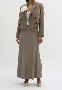 Giacca marrone doppiopetto con un'accoppiatura di fiori bianchi, abbinata a una lunga gonna beige. L'outfit presenta una texture liscia e un design aderente.