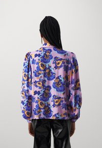 Blusa floral en púrpura, con patrones florales en azul y amarillo, mangas abullonadas y un tejido suave y liso. Pantalones negros brillantes visibles.
