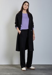 Abrigo negro largo sobre una blusa de color púrpura claro, combinado con pantalones negros y zapatillas blancas. Presenta un diseño ajustado y una textura suave.