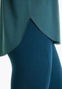 Haut de sport teal avec un ourlet incurvé, associé à un legging teal uni. Les deux vêtements présentent des matériaux lisses et extensibles ainsi qu'une coupe ajustée.