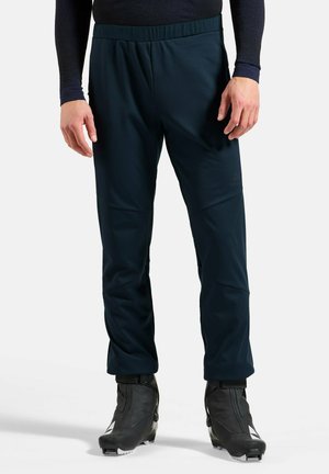Person wearing navy blue slim-fit pants and black ski boots standing against a neutral background.