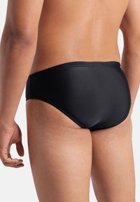 Bañador tipo slip negro fabricado con material liso y elástico, con un diseño de tiro bajo y cobertura mínima en la parte trasera y los laterales.