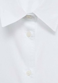 Chemise blanche à col pointu avec trois boutons blancs le long de la patte de boutonnage avant, montrant des détails de couture.