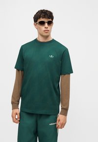 Άνδρας που φοράει σκούρο πράσινο φόρμα Adidas με καφέ μακριά μανίκια από κάτω και ανοιχτό καφέ γυαλιά ηλίου, στέκεται μπροστά σε λευκό φόντο.