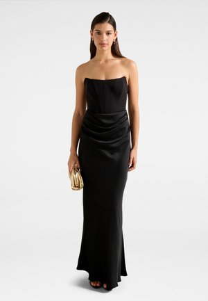 Vrouw draagt een strapless zwarte avondjurk en houdt een kleine glanzende gouden clutch vast tegen een eenvoudige lichte achtergrond.