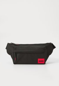 Sacoche en tissu noir avec poche zippée avant, sangle ajustable et patch du logo "HUGO" rouge dans le coin inférieur droit.