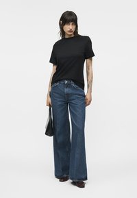 Femme portant un haut noir à manches courtes, jean bleu taille haute à jambes larges, tenant un sac à main noir, debout devant un fond blanc.