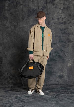 Beige takki, jossa on etutaskut ja ruskea hattu vihreän paidan päällä. Malli pitää mustaa, soikeaa laukkua, jossa on oranssi logo. Ruskeat kengät viimeistelevät asusteet.