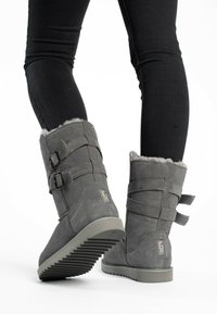 Botas de tobillo de ante gris con ribete de pelo sintético, que presentan dos hebillas laterales y una suela de goma texturizada. Mostradas con jeans ajustados negros.