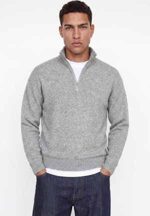 Homme portant un pull gris clair à zip quart de fermeture sur une chemise blanche avec un jean bleu foncé, debout les mains dans les poches devant un fond blanc.