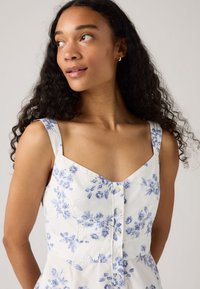 Giovane donna con lunghi capelli ricci che indossa un vestito bianco con un motivo floreale blu e dettagli di bottoni, che guarda di lato.