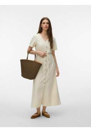 Hosszú barna hajú nő krémszínű, gombos midi ruhában, kezében nagy barna és fekete csíkos bevásárlótáskával, barna lapos cipőben.