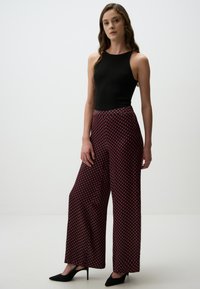 Zwarte mouwloze top met hoge hals, gecombineerd met wijde broek met een rood-wit geometrisch patroon. Zwarte puntige hakken.