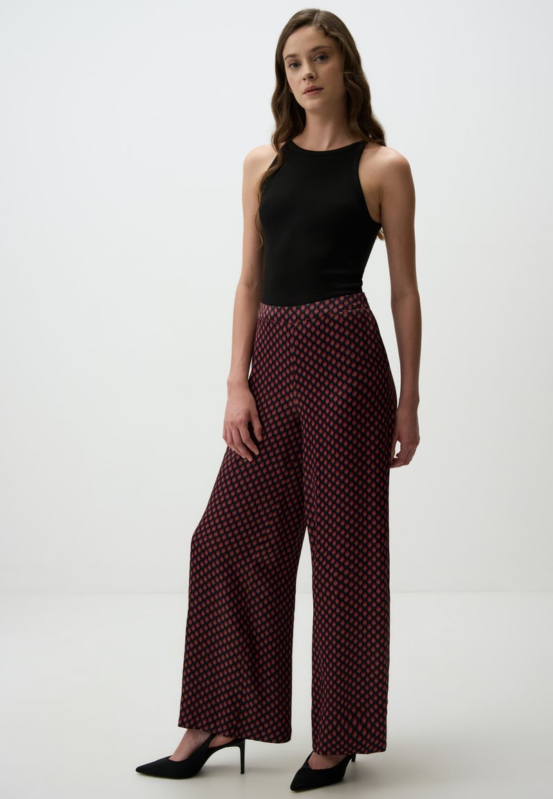 Zwarte mouwloze top met hoge hals, gecombineerd met wijde broek met een rood-wit geometrisch patroon. Zwarte puntige hakken.