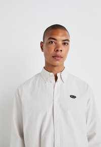 Camicia bianca con colletto e sottili righe verticali, con logo sul lato sinistro del petto, realizzata in tessuto leggero.