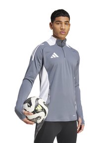 Giacca sportiva grigia con accenti bianchi, collare con mezza zip, maniche lunghe e fori per il pollice, abbinata a un pallone da calcio bianco e nero.