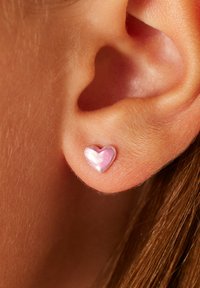 Pinke, herzförmige Ohrringe aus glänzendem Material, eingearbeitet in einen silberfarbenen Hintergrund. Aufnahmedetail eines menschlichen Ohrs.