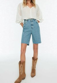 Mujer con blusa blanca transparente, bermudas de mezclilla azul de cintura alta y botas vaqueras color canela hasta la mitad de la pantorrilla, de pie con las manos en los bolsillos.