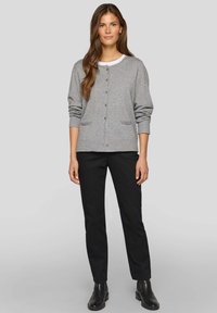 Graue Strickjacke mit Knopfverschluss vorne, gerippten Bündchen und zwei Vordertaschen. Kombiniert mit schwarzen Hosen und Stiefeletten.