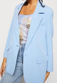 Ljusblå blazer i mjukt tyg med slag, enkel knäppning och framfickor, lager på en mönstrad topp.