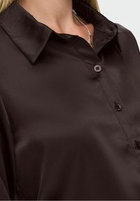 Gros plan sur une personne portant une chemise en satin marron foncé à boutons avec un col et un délicat collier en argent.