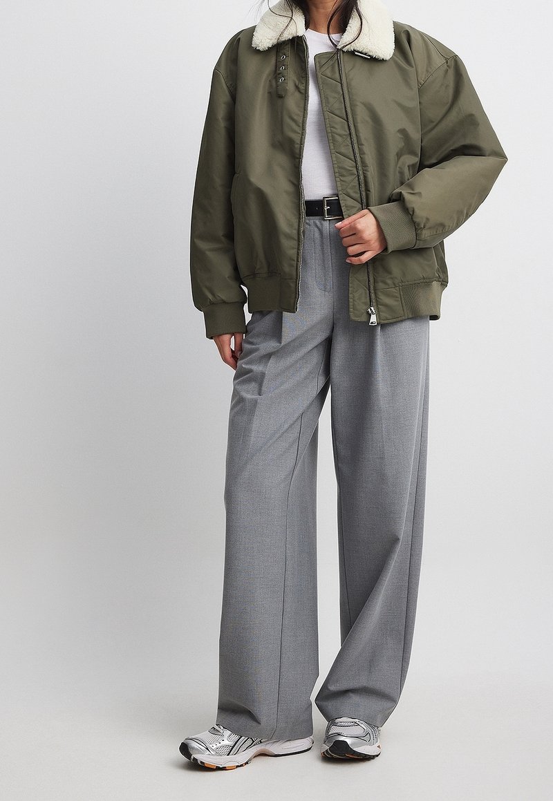 Cazadora bomber verde oliva con cuello de borrego sintético, combinada con pantalones de pierna ancha gris claro y zapatillas deportivas plateadas.