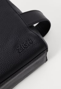 Black leather bag with a textured surface, featuring a prominent logo, a smooth strap, and minimalistic design.