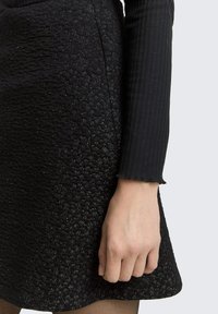 A jobb kéz egy fekete, textúrás szoknya mellett pihen, amelyen finom fényes részletek találhatóak. Hosszú ujjú fekete bordázott felsőt visel, lágyan fodros mandzsettával.