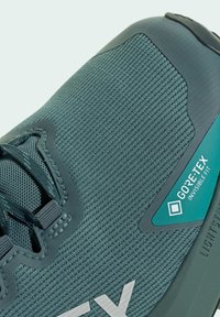 Scarpa da corsa verde scuro con tomaia in mesh testurizzato, zona lacci rinforzata e etichetta GORE-TEX. Profilo snello con suola in gomma.