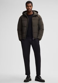 Giacca a vento verde scuro con cappuccio, con chiusura a zip e design trapuntato. Abbinata a pantaloni neri e scarpe nere con lacci.