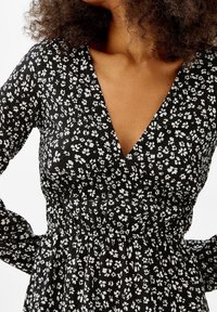 Kvinna i svart klänning med vitt blommönster och djup V-ringning, synlig från axlarna till midjan, med lockigt hår delvis synligt.