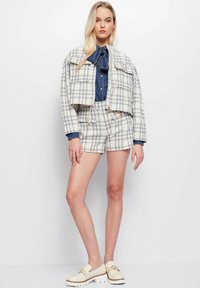 Witte en blauwe geruite cropped jas met twee voorzakken, gecombineerd met bijpassende shorts. Daaronder een denimshirt met een strikdetail. Crème schoenen.