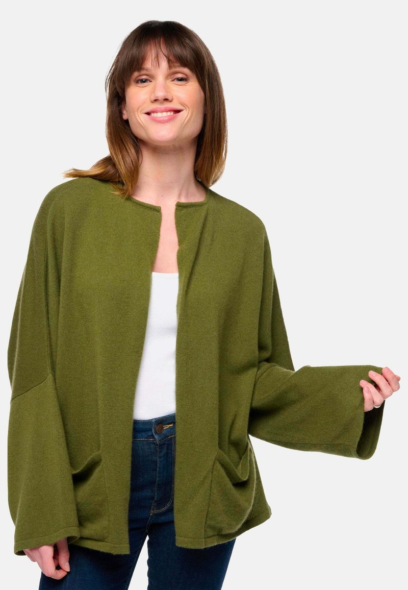 Cardigan vert surdimensionné en tissu doux, avec un col rond, des manches larges et des poches avant. Stylé par-dessus un haut blanc et un jean.