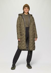 Gequilted, olijfgroen lange jas met een capuchon, voorzien van zijzakken en een ritsluiting. Gedragen over donkere broeken en zwarte chunky laarzen.