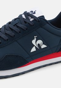 Sneaker blu navy con una combinazione di materiali lisci e testurizzati. Logo bianco sul lato, dettagli rossi sulla suola.
