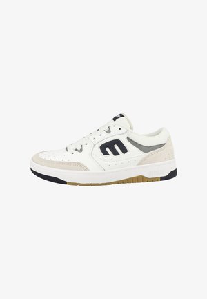 White sneaker with beige accents and black logo. Features perforated texture, flat laces, and a sturdy sole with a gum rubber bottom.