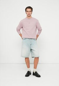 Világos rózsaszín hosszú ujjú ing logóval, világoskék farmer rövidnadrág, fehér zokni és fekete gumipapucs. A modell kezeit csípőjére téve áll.