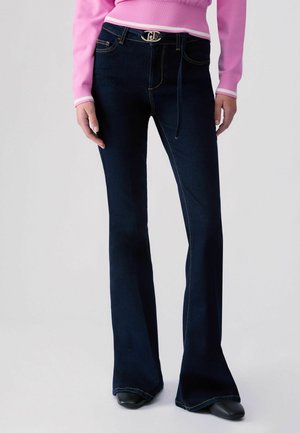 Donna con jeans a zampa d’elefante blu scuro, cintura nera e maglietta rosa a maniche lunghe, in piedi con le mani rilassate lungo i fianchi.